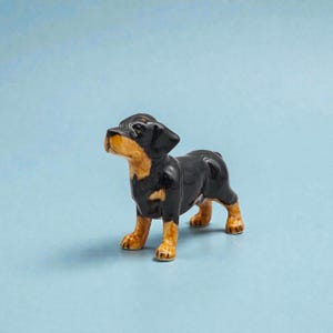 Puede incluir: Figura de cerámica de un cachorro, con pelaje negro y marcas marrones en la cara y las patas. El cachorro está de pie sobre sus cuatro patas, con la cabeza levantada. El fondo es azul claro liso.