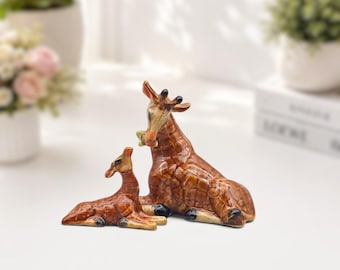 May include: A pair of ceramic giraffe figurines featuring a glossy brown and tan crackle glaze finish. The larger giraffe is in a seated position, while the smaller giraffe is lying down beside it, both displaying detailed facial features.