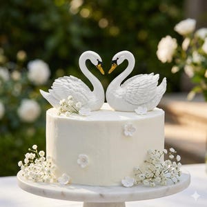 May include: A white wedding cake with two swan cake toppers facing each other. The swans have yellow beaks. The cake is decorated with white flowers and sits on a marble cake stand. The background is blurred with greenery and white flowers.