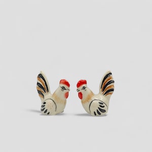 May include: Two ceramic rooster figurines. Each rooster has a red comb and wattle, with a cream-colored body and black and brown tail feathers. The roosters are facing each other, with a white background.