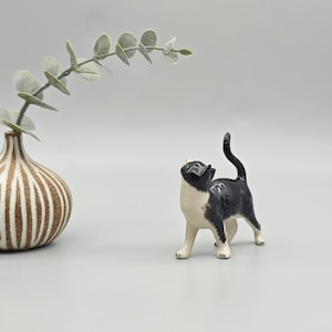 May include: A ceramic figurine of a black and white cat with its tail up, looking upwards. The cat is standing on all fours. A decorative vase with brown and white stripes and a sprig of green leaves are in the background.