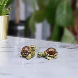 May include: Two small ceramic turtle figurines featuring glossy green bodies and textured brown shells. One turtle is positioned in an upright sitting posture, while the other is depicted in a crawling position with its head extended forward.