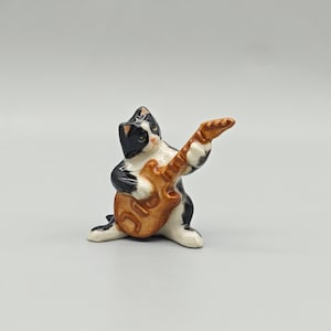 Puede incluir: Una figura de cerámica de un gato blanco y negro tocando una guitarra marrón. El gato está en posición agachada, con las patas en el suelo y la cabeza ligeramente inclinada. La guitarra es de color marrón claro.
