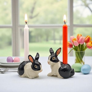 Puede incluir: Dos candelabros de cerámica con forma de conejito, uno con una vela blanca y otro con una vela roja. Los conejos son blancos y negros con orejas rosas y ojos amarillos. Un jarrón de tulipanes y huevos de Pascua se encuentran en el fondo.