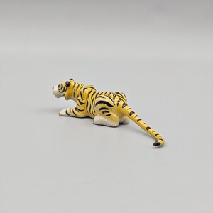May include: A small, yellow and black tiger figurine in a resting pose. The tiger has black stripes and white paws. The figurine is on a plain, light gray background. The tiger's tail is extended.
