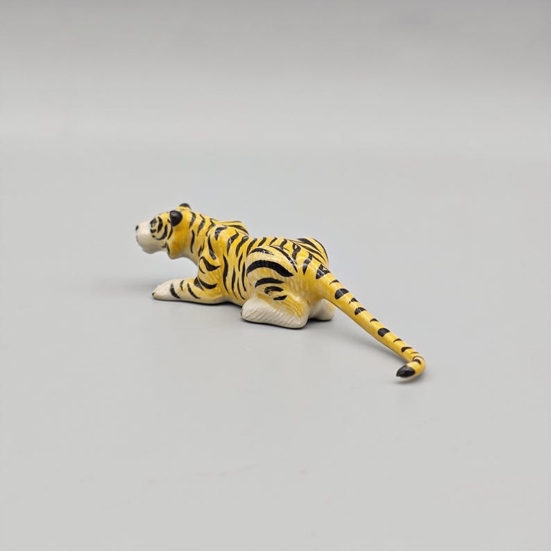 May include: A small, yellow and black tiger figurine in a resting pose. The tiger has black stripes and white paws. The figurine is on a plain, light gray background. The tiger's tail is extended.