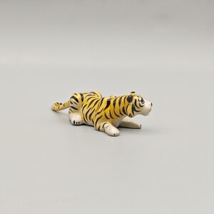 May include: A ceramic tiger figurine in a resting pose. The tiger has a yellow body with black stripes and white paws and face. The figurine is on a gray background.