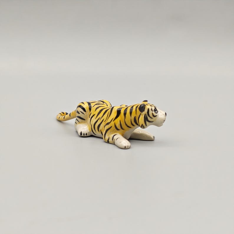 May include: A ceramic tiger figurine in a resting pose. The tiger has a yellow body with black stripes and white paws and face. The figurine is on a gray background.