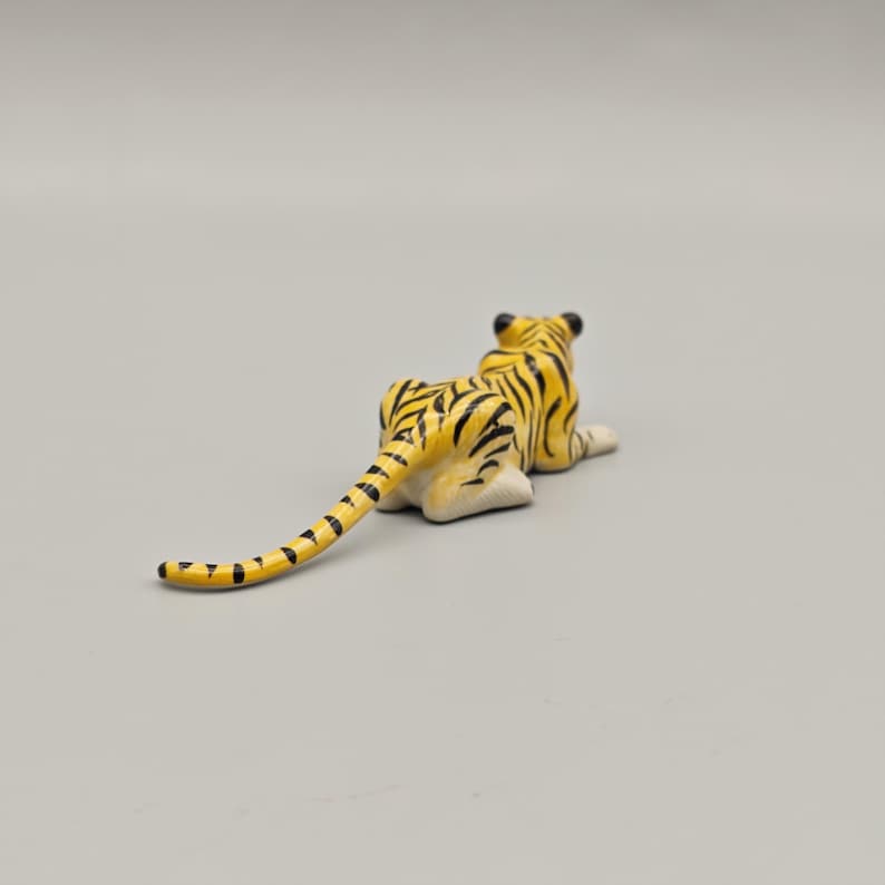 May include: A small, ceramic tiger figurine in a resting pose. The tiger has a yellow body with black stripes and a long tail. The figurine is on a plain gray background.