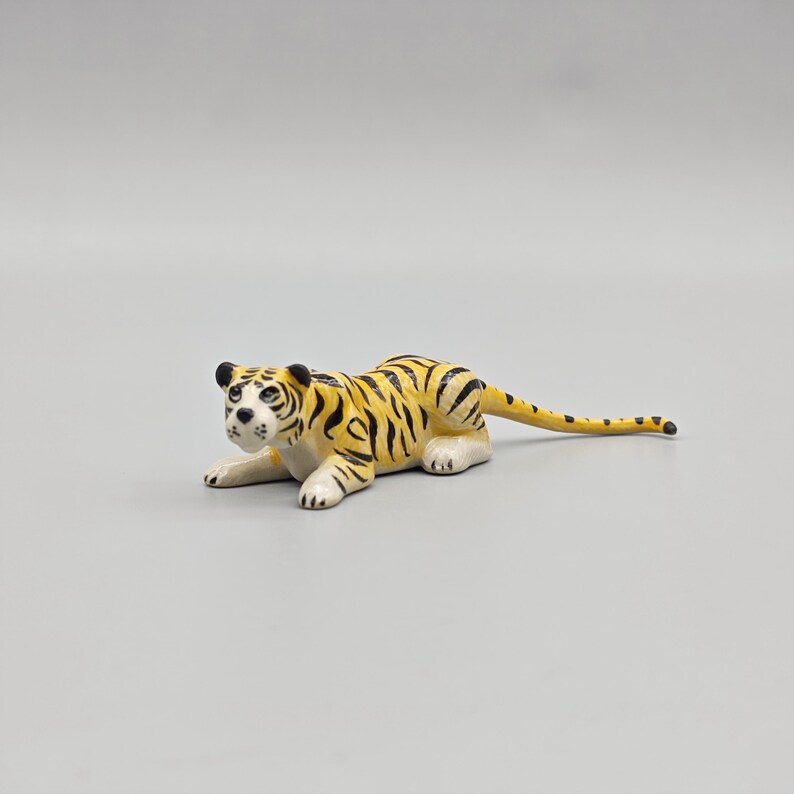 May include: A small, yellow and black tiger figurine in a resting pose. The tiger has black stripes, white paws, and a white face with black markings. The figurine is lying down on a gray surface.