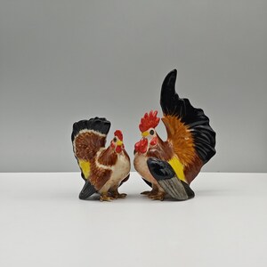 May include: A pair of ceramic rooster figurines. The rooster on the left is brown, black, and yellow with a red comb. The rooster on the right is brown, black, and yellow with a red comb and a black tail.