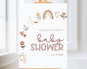 May include: A white baby shower sign on a wooden easel. The sign features watercolor illustrations of a rainbow, unicorn, flowers, and hearts. The text reads "Welcome to Ashley's baby shower" and the date "June 18, 20XX".