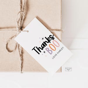 May include: A gift tag with the words "Thanks BOO Love Harley" in black, pink, and purple. The tag is attached to a wrapped package with twine. The background is a light beige colour.