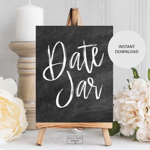 Pode incluir: Um sinal de quadro-negro preto com texto branco que diz "Date Jar" em um cavalete de madeira. O sinal é cercado por flores brancas e uma vela.