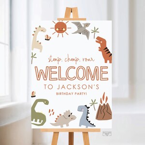 Könnte beinhalten: Ein weißes Willkommensschild auf einer hölzernen Staffelei mit Dinosaurier-Motiv. Das Schild zeigt Illustrationen von Dinosauriern, einer Sonne und Palmen mit dem Text "stomp, chomp, roar WELCOME TO JACKSON'S BIRTHDAY PARTY!"