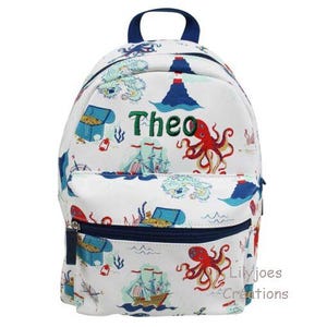 Könnte beinhalten: Weißer Rucksack mit nautischem Thema in Blau, Rot und Grün. Der Rucksack hat eine Reißverschlusstasche und zeigt einen Oktopus, ein Schiff und eine Schatztruhe. Der Name "Theo" ist auf der Vorderseite des Rucksacks gestickt.