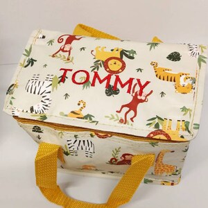 May include: Cream-coloured lunch bag with a jungle animal pattern, featuring zebras, lions, tigers, monkeys, and giraffes. The bag has yellow handles and the name "TOMMY" embroidered in red. The bag is rectangular in shape.