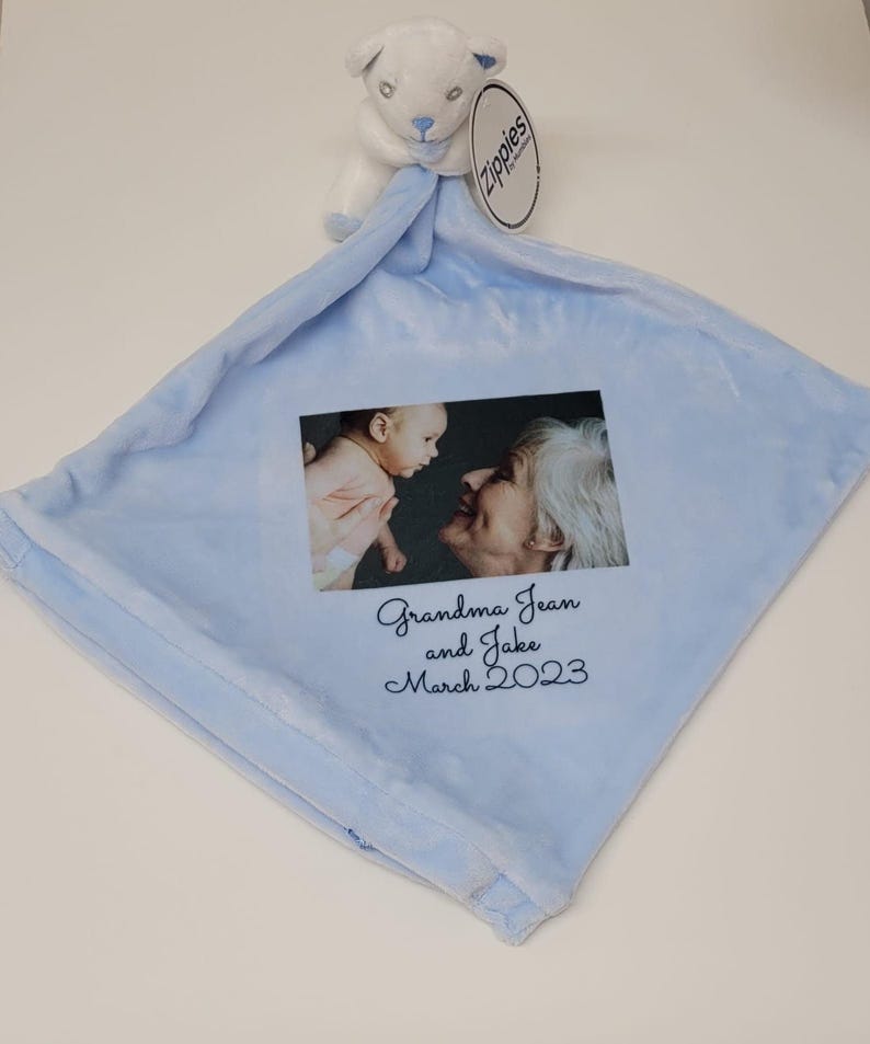 Peut inclure: Une couverture pour b&eacute;b&eacute; bleu clair avec un animal en peluche blanc attach&eacute;. La couverture pr&eacute;sente une photo d'une grand-m&egrave;re et d'un b&eacute;b&eacute; avec le texte "Grandma Jean and Jake March 2023" imprim&eacute; dessus.