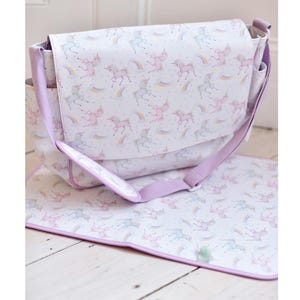 May include: A white diaper bag with a flap closure and a matching changing pad. The bag and pad feature a repeating pattern of pastel-colored unicorns and rainbows. The bag has a lavender shoulder strap and trim.