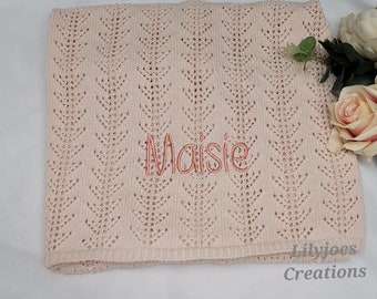 May include: A pink crocheted baby blanket with a delicate pattern and the name "Maisie" embroidered in a light pink color. The blanket is folded and laid on a white surface.