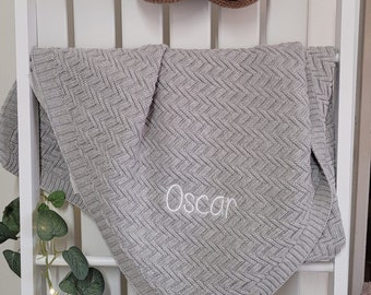 May include: A gray knitted baby blanket with the name "Oscar" embroidered on it. The blanket is folded and draped over a white ladder. A crocheted sloth toy hangs above the blanket.