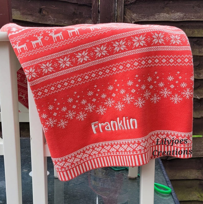 May include: Red and white knitted baby blanket with a snowflake and reindeer pattern. The blanket has the name "Franklin" embroidered on it. The blanket is made by Lilyjoes Creations.
