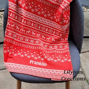 May include: Red and white knitted throw blanket with a traditional Christmas pattern featuring snowflakes, trees, and reindeer. The blanket has the name "Franklin" embroidered in white on the bottom.