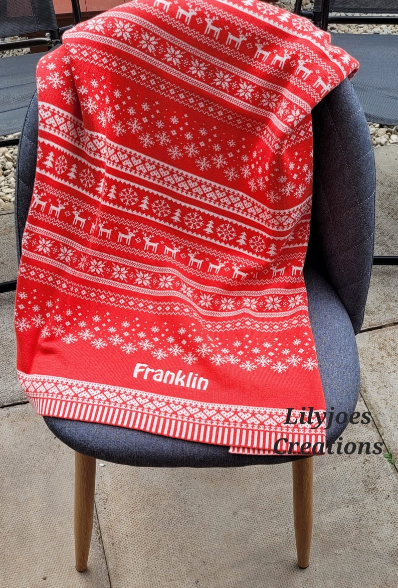 May include: Red and white knitted throw blanket with a traditional Christmas pattern featuring snowflakes, trees, and reindeer. The blanket has the name "Franklin" embroidered in white on the bottom.