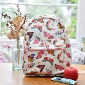 Op de afbeelding: Witte rugzak met een patroon van kleurrijke vlinders en een roze rits. De rugzak staat op een houten tafel naast een rode appel en een snackreep. Een vaas met gedroogde bloemen staat op de achtergrond.