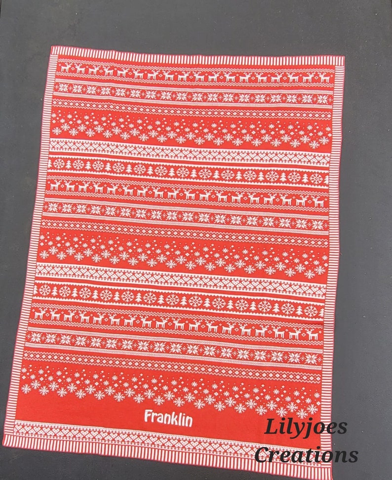 May include: Red and white Christmas themed kitchen towel with a Scandinavian pattern of snowflakes, reindeer, and trees. The towel has the name "Franklin" and the text "Lilyjoes Creations" printed on it.