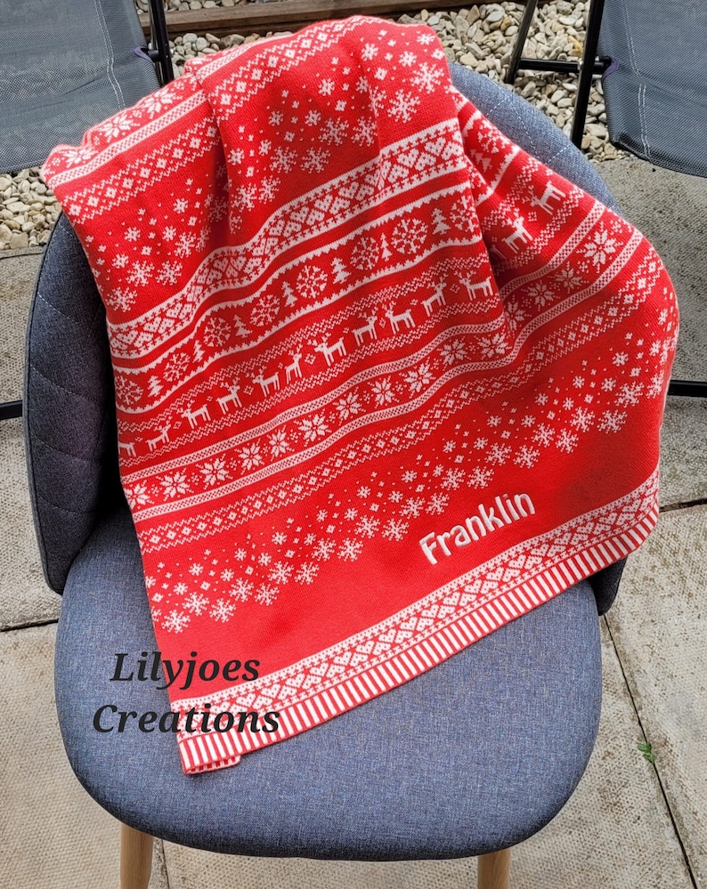 May include: Red and white knitted throw blanket with a snowflake and reindeer pattern. The blanket has the name "Franklin" embroidered on it.