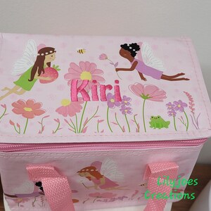 May include: Pink insulated lunch bag with a fairy and floral design. The bag has a zip closure, pink handles, and the name "Kiri" embroidered in pink. The design includes fairies, flowers, a bee, and a frog.