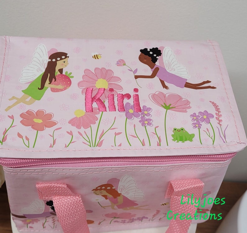May include: Pink insulated lunch bag with a fairy and floral design. The bag has a zip closure, pink handles, and the name "Kiri" embroidered in pink. The design includes fairies, flowers, a bee, and a frog.
