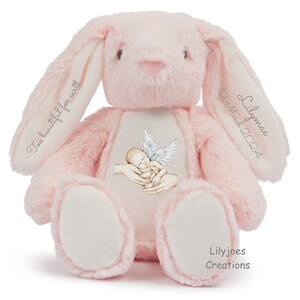 Op de afbeelding: Een roze pluche konijn met geborduurde tekst "Too beautiful for earth" op het ene oor en "Lilymae 12 March 2024" op het andere oor. Het konijn heeft een witte vlek op zijn borst met een geborduurd beeld van een babyengel met vleugels en handen die de baby vasthouden. De voeten van het konijn zijn wit en de tekst "Lilyjoes Creations" is geborduurd op de onderkant van een voet.