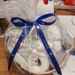 May include: A gray and white baby gift basket with a plush elephant toy, a blanket, and a rattle. The basket is wrapped in clear cellophane and tied with a blue ribbon with gold stars and the name "CHASE" printed on it.