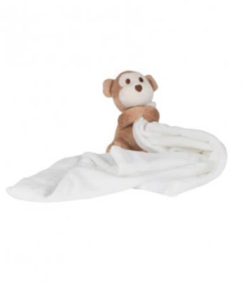 Peut inclure: Une couverture en peluche blanche avec un singe en peluche marron attach&eacute;. Le singe tient la couverture avec ses bras.
