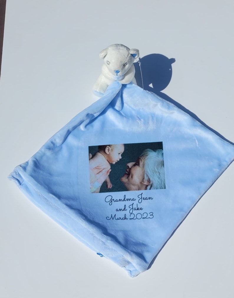 Peut inclure: Une couverture de b&eacute;b&eacute; bleue avec un animal en peluche blanc attach&eacute;. La couverture pr&eacute;sente une photo d'un b&eacute;b&eacute; et d'une femme &acirc;g&eacute;e avec le texte "Grandma Jean and Jake March 2023" imprim&eacute; dessus.