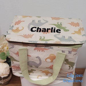 May include: A white lunch bag with a dinosaur print and a green zipper. The bag has a green handle and the name "Charlie" is embroidered on the front.