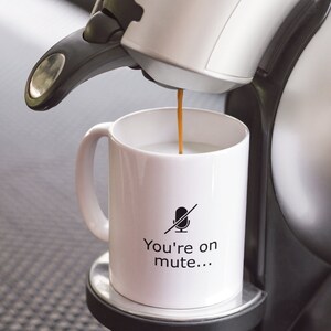 May include: A white coffee mug with a black microphone with a line through it and the text "You're on mute..." printed on it. Coffee is being poured into the mug from a white coffee maker.