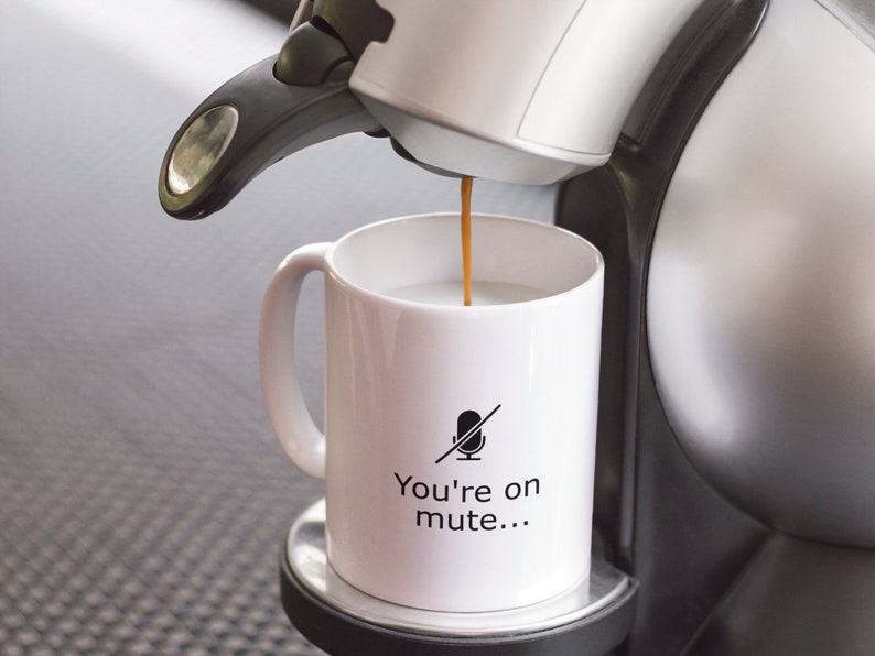May include: A white coffee mug with a black microphone with a line through it and the text "You're on mute..." printed on it. Coffee is being poured into the mug from a white coffee maker.