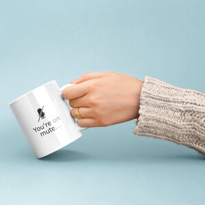 May include: White ceramic mug with a black microphone icon and the text "You're on mute..." printed on the side. The mug is being held by a hand wearing a beige sweater.