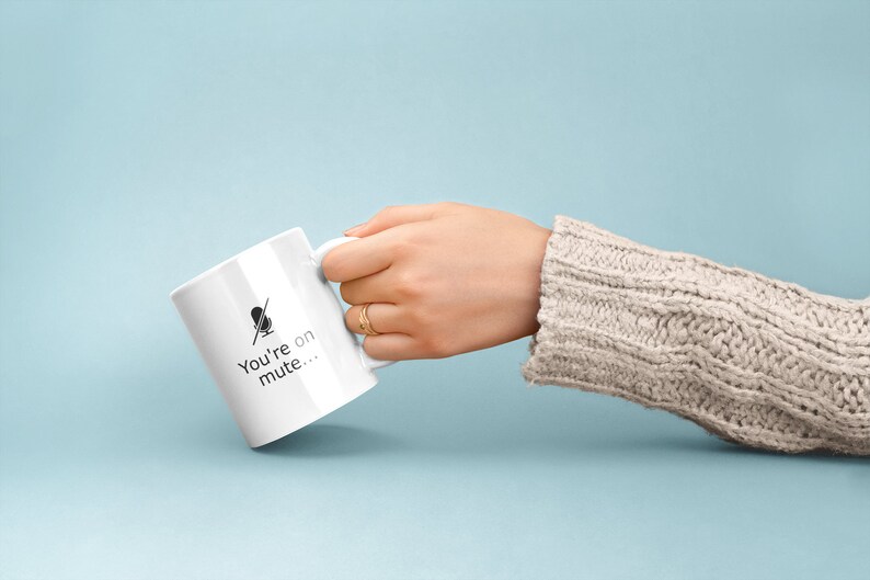 May include: White ceramic mug with a black microphone icon and the text "You're on mute..." printed on the side. The mug is being held by a hand wearing a beige sweater.