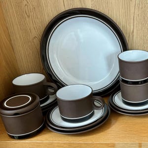 May include: A brown and white ceramic dish set. The set includes a large plate, cups, saucers, a creamer, and a sugar bowl. The cups and sugar bowl have a black stripe. The set is displayed on a wooden shelf.