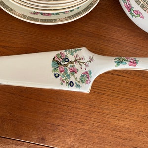 May include: A white ceramic cake server with a gold trim and a floral design featuring a tree with pink flowers and green leaves.