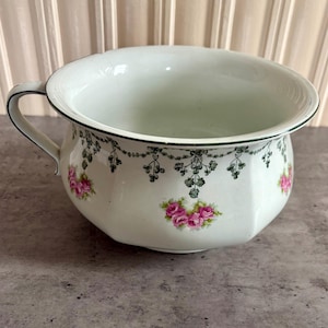 May include: A white ceramic chamber pot featuring a handle and a decorative pattern of pink roses and green foliage swags around the exterior. The rim is accented with a thin green line, and the body has a slightly faceted, rounded shape.