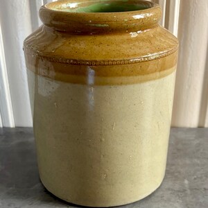 May include: A cylindrical stoneware crock featuring a two-tone glaze with a light beige lower body and a speckled brown upper section. The rim is thick and rounded, revealing a light green interior glaze and a decorative embossed band.