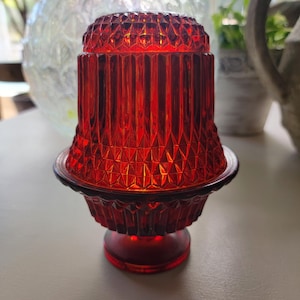 May include: A ruby red glass candle holder with a geometric pattern. The candle holder has a pedestal base and a flared top. The glass is translucent, allowing light to pass through. The candle holder is a decorative item.
