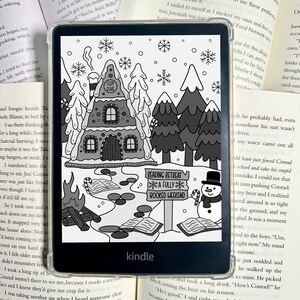May include: Kindle e-reader with a black and white winter scene on the screen. The image features a gingerbread house, trees, snowflakes, and a snowman holding a candy cane. Text on the screen reads "Cozy Cove" and "Reading Retreat A Fully Booked Weekend."