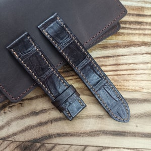 May include: Two brown leather watch straps with a crocodile pattern and orange stitching. The straps are laid out on a wooden surface.