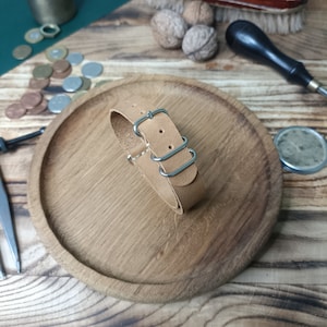 Può includere: Un cinturino da orologio in pelle marrone chiaro con fibbia argentata e tre passanti. Il cinturino è su una superficie di legno.